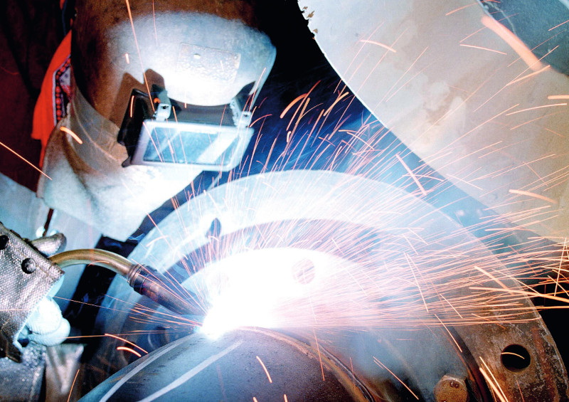 Foto van een lasser in beschermende kleding met laskap op bij metaal waarbij veel vonken zichtbaar zijn.